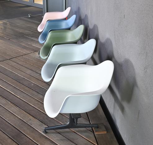 Das Bild zeigt eine Reihe von fünf modernen Stühlen mit einem einzigartigen, geschwungenen Design, die vor einer grauen Wand auf einer Holzterrasse stehen. Die Stühle sind gestaffelt angeordnet, wobei jeder Stuhl den dahinter stehenden leicht überlappt. Die Farben der Stühle, von vorne nach hinten, sind weiß, hellblau, grün, blau und rosa. Jeder Stuhl hat eine glatte, matte Oberfläche und eine markante, ergonomische Form mit einer breiten Sitzfläche und integrierten Armlehnen. Die Stühle werden von einem schwarzen Metallfuß mit einem fünfzackigen Stern getragen, der für Stabilität sorgt. Das Holzdeck hat eine natürliche, dunkelbraune Farbe mit sichtbarer Holzmaserung, und die graue Wand hat eine strukturierte Oberfläche. Die Beleuchtung auf dem Bild lässt vermuten, dass es Tag ist, und die Schatten, die die Stühle auf die Wand werfen, deuten auf eine Lichtquelle auf der linken Seite hin.