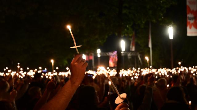 Im Vordergrund zwei dünne brennende Kerzen mit rundem Schutz gegen herunterfließendes heißes Wachs. Im Hintergrund viele weitere brennende Kerzen und Lampen sowie Bäume in der Dunkelheit des Abends.