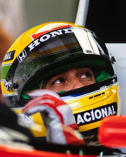 A racing driver is seated in a car, wearing a yellow helmet with brand logos on it. The visor is slightly open, revealing the driver's eyes looking upwards and to their left.