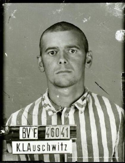 A fragment of a mugshot registration photograph from Auschwitz. A man with a shaved head wearing a striped uniform photographed (front with bare head). The prisoner number is visible on a marking board on the left.