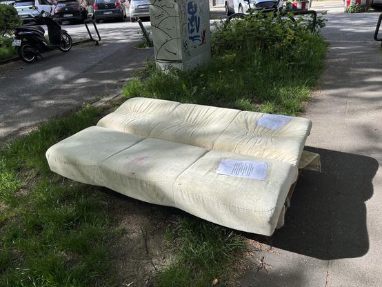 Das Bild zeigt ein altes, beige Sofa, das auf dem Bürgersteig vor einem Straßenelement mit Graffiti abgestellt ist. Das Sofa ist zerknittert und zeigt deutliche Gebrauchsspuren, einschließlich einer roten Fleckenmarkierung. Auf dem Sofa liegen zwei Papiere, die möglicherweise Informationen oder Anweisungen enthalten. Im Hintergrund sind geparkte Autos und ein Motorrad zu sehen, sowie ein Straßenelement mit Graffiti, das teilweise die Buchstaben "EV" zeigt. Die Umgebung wirkt urban, mit Gras und Pflanzen zwischen dem Bürgersteig und der Straße.