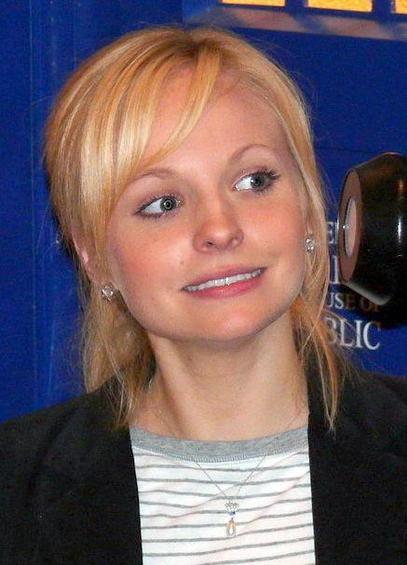Georgia Moffet, portant un collier avec un pendentif en forme de goutte, est vêtue d'une chemise à rayures grises et blanches sous une veste noire. Elle se tient devant une cabine téléphonique bleue avec l'inscription 'PUBLIC' visible en arrière-plan. La lumière met en valeur ses traits et l'ambiance générale est lumineuse et chaleureuse.