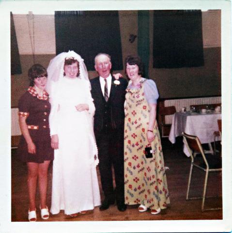 A scan of a square, colour photo, taken in the early 1970s. It's in a hall, possibly one belonging to a school, with wooden flooring and windows high up on the walls in the background, framed by dark green curtains. To the right there are tables pushed together in rows with tablecloths overs them and place settings for food and drink, along with small, padded chairs. To the left of the view, and the main focus, there are four people. A woman stands in her bridal dress with veil pulled back from her smiling face. An older man in dark suit and tie and with a flower in his buttonhole is to the right of her. Either side of them are two other women, both dark-haired, both wearing odd choices of clothing. The woman on the left has a short-sleeved dress to her thighs in a burgundy-brown colour with red and white chequerboard patterns on the sleeve tips, collar, and pocket tops. The woman on the right is wearing a floor-length pale yellow dress with abstract mosaic flower designs on it. Because it has thin shoulder straps she's wearing this over a pale blue blouse and is carrying a compact camera in her hand.