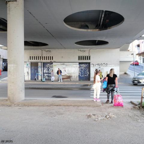 Farbfoto, das eine Bushaltestelle unter einer Gebäudebrücke in einem eher heruntergekommenen Stadtteil von Rom zeigt. Die Gebäude im Stil des Brutalismus sind deutlich lädiert, Wände und Türen sind mit Grafitti verschönert. An der Haltestelle warten zwei Leute auf den Bus, Tochter und Mutter mit Gepäck weiter vorn sind gerade dabei, die Straße zu überqueren.
