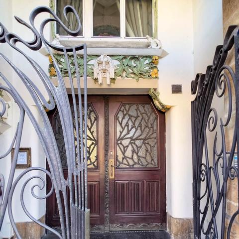 A detailed view through an ornate wrought iron gate towards the entrance of Hilbert House in Prague. The warm-toned wooden double doors feature stained glass panels with organic, plant-like designs. Above the doorway, an elaborate Art Nouveau lintel in shades of cream, green, and gold includes sculpted organic forms and a stylized mask-like face. A window with white frames and light curtains is visible above the lintel. The building's facade is a muted cream color.