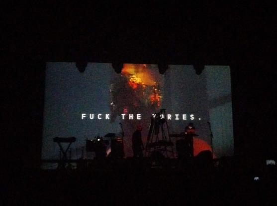 Test Dept. no palco. No ecrã atrás deles a mensagem é "Fuck the Tories."