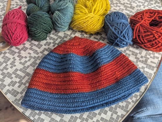Mütze aus roter und blauer Wolle auf Tisch, im Hintergrund weitere Wolle in verschiedenen Farben