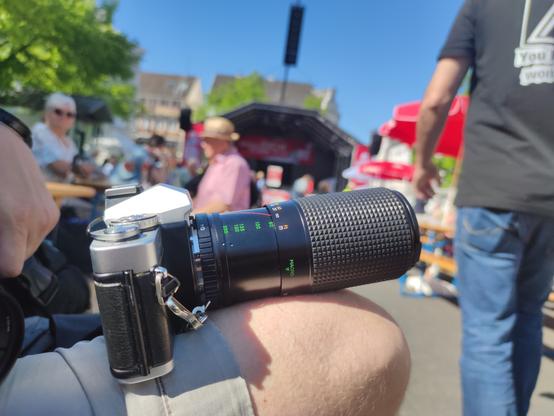 Eine Minolta XD7 mit Travenar 80-200mm f/4.5 auf der Mainveranstaltung in Siegburg. die Kamera ruht auf meinem Knie, im Hintergrund die Bühne. Rechts läuft eine blaue Jeans ins Bild.