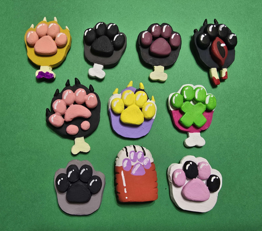 10 different polymer clay paw pins, from top  left to bottom right: yellow with pink pads and purple blood on bone, grey brown with black pads white bone, black with flesh pink pads and bone, light black with black toe pads, an eye for the palm and wrist bones with blood, ferret shaped black with pink pads and bone, purple with white and darker purple toes and yellow pads/claws, pink with white toes green pads x shaped palm and bone, light gray with black pads, pooltoy paw with orange fur and white toes purple pads, and finally white with black and pink pads