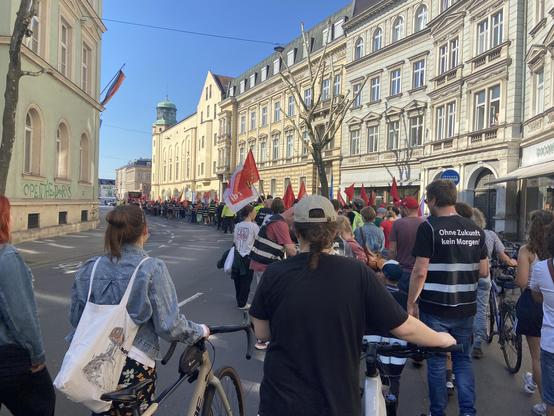 Bild einer Demo zum 1. Mai. Es sind hunderte Menschen in einer Kurve von hinten fotografiert. Die Menschen tragen Fahnen und teilweise Warnwesten.

Im Hintergrund ist eine Häuserzeile und blauer Himmel zu sehen.