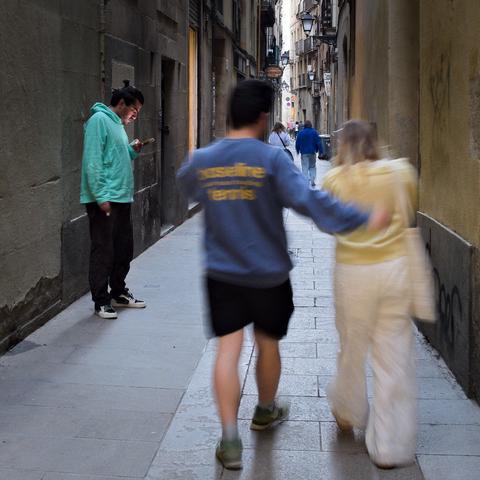Gasse mit Fußgängern. Ein unscharfes Pärchen rechts im Vordergrund. Links dahinter eine scharf abgebildete Person, die auf ihr Handy guckt.