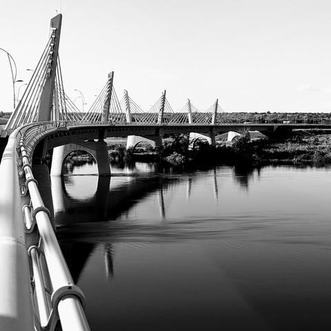 Black and white photo of the Kazungula Bridge which spans between Botswana and Zambia. The photo is taken at an angle which reveals the curvature of the bridge, the cable-stayed design and the expanse of the river beneath. At the far end Zambia is visible. The bridge took 10 years to build and was opened in 2021. The bridge is at the crux between 4 countries' borders: Botswana, Zimbabwe, Zambia, and Namibia.