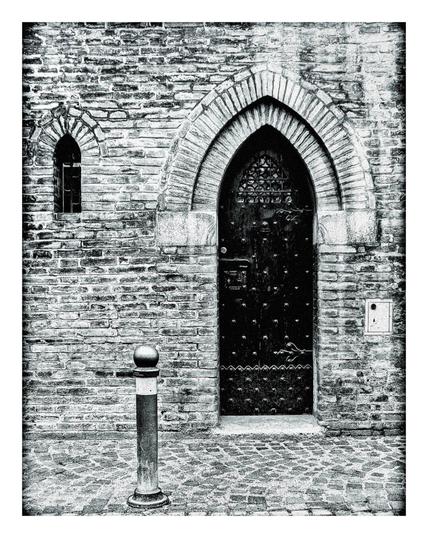 A black and white photo of the wall of a building from a street. On the left hand side there is a small arched window. On the right hand side an vaulted door with studs on it. in the foreground between the window and door is a small bollard at street level.