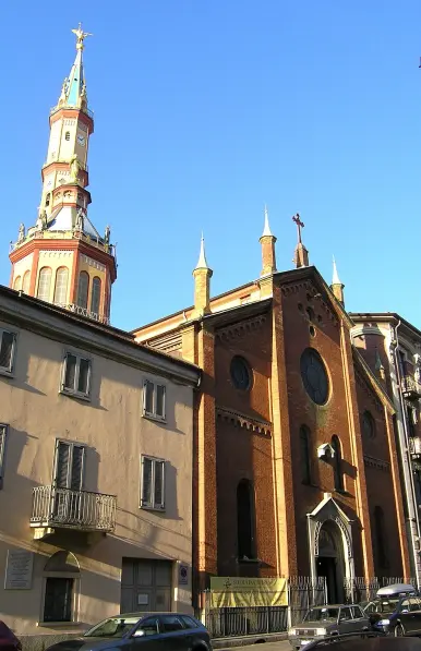 The image features a building with a brick facade, likely a church, and a distinctive tower with a spire. The tower appears to be ornate, with colorful elements and a cross at its top. The architecture suggests a historical style, and the setting seems to be in an urban area with other buildings nearby. The sky is clear, indicating a sunny day.

Image Credits: Wikimedia / Marco Plassio / CC BY-SA 3.0