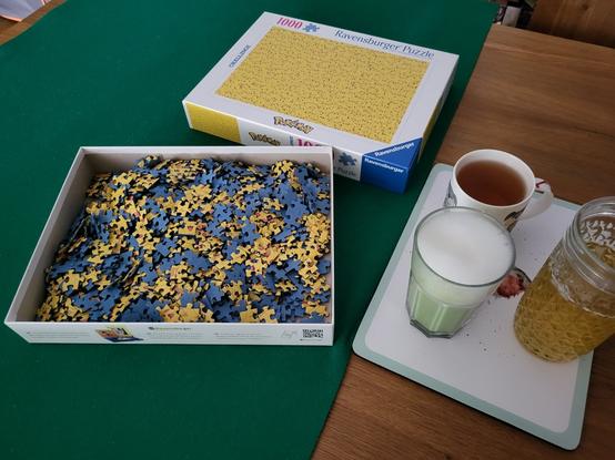Ein 1000-teiliges Pokémon-Puzzle mit von Ravensburger liegt bereit zum Start. Das Motiv sind zig gelbe Pikachus. 
Daneben stehen drei Getränke: ein schwarzer Tee, ein grünes Getränk mit Schaum (Matcha Latte), und ein hellgelber Grüner in einem Glas