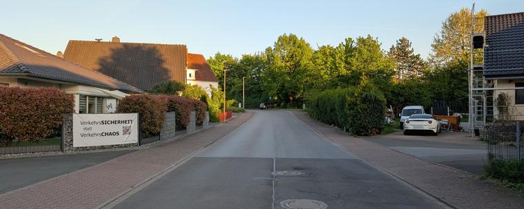 Eine Straße in einem Wohngebiet. Rechts und links der breiten Fahrbahn, auf der man sehr deutlich die vier Spuren der die Straße nutzenden Fahrzeuge sieht, sind Gehwege mit abgesenkten Bordsteinen angelegt. Links an einem Gabionenzaun hängt ein Plakat mit der Aufschrift "VerkehrsSICHERHEIT statt VerkehrsCHAOS". Auf der Fahrbahn ist kein Fahrzeug zu sehen.