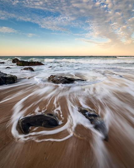 a vertical seascape showing a receding wave weaving around and between the rocks. The receding wave makes white streaks against the brown sand. There are some nice high clouds in the sky that have not yet turned red.