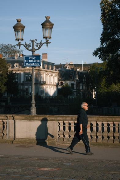 A man walking briskly across a bridge, past a sign reading "Pont du Théâtre (Theaterbruck)". His shadow is cast on one of the bridge's walls.