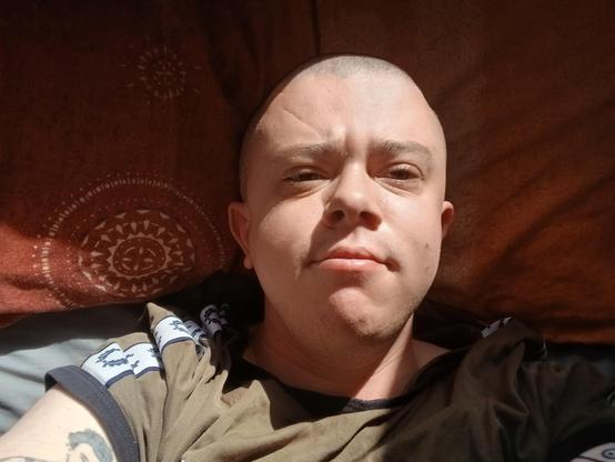 A white trans mask wearing a green top lying in bed. He has a shaved head and the sun is on his face