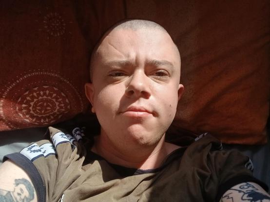A white trans mask wearing a green top lying in bed. He has a shaved head and the sun is on his face