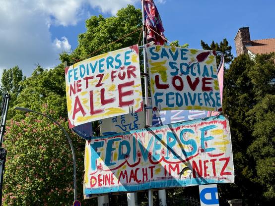A collection of colorful, hand-painted banners promoting the "Fediverse." Messages include "Netz für Alle," "Save Social Love," and "Deine Macht im Netz." The background features trees and a blue sky.