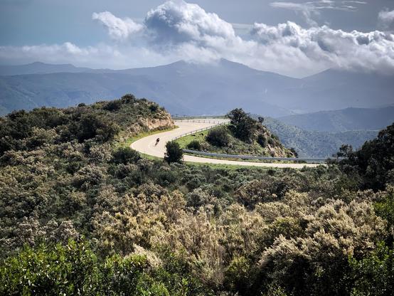 Une route avec une cycliste  serpente dans un paysage vallonné avec des montagnes et quelques nuages dans le fond.