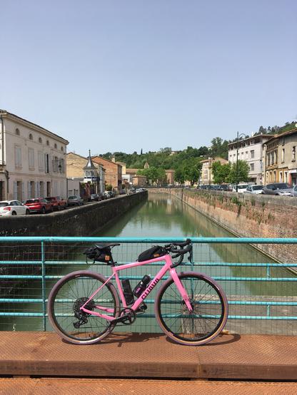 Posé contre le parapet en grille métallique d'un des ponts du Canal Latéral à la Garonne à Moissac, un vélo de gravel Guava Spot rose et violet.