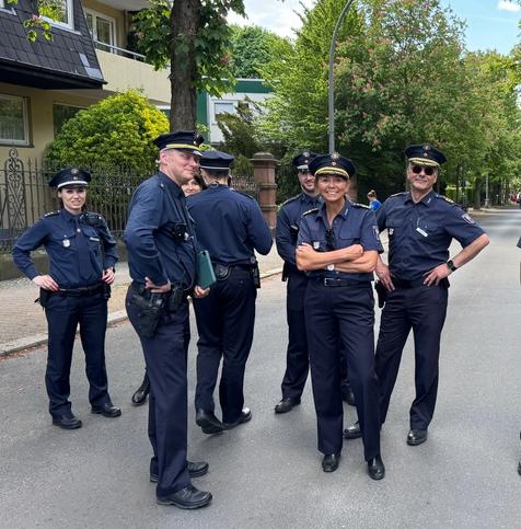 A group of six police officers in uniform stand on a street, smiling and interacting. They are dressed in dark blue uniforms with badges, and there are trees and buildings in the background.