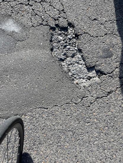 An irregularly shaped hole in an asphalt surface, part of bike wheel visible.