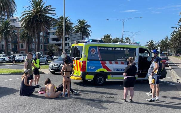 An ambulance is on a road.  In the foreground is a group of people including a person wearing a cycling helmet lying on the ground, an ambo, two people in cycling clothes, two women in bikinis.   Palm trees in background.