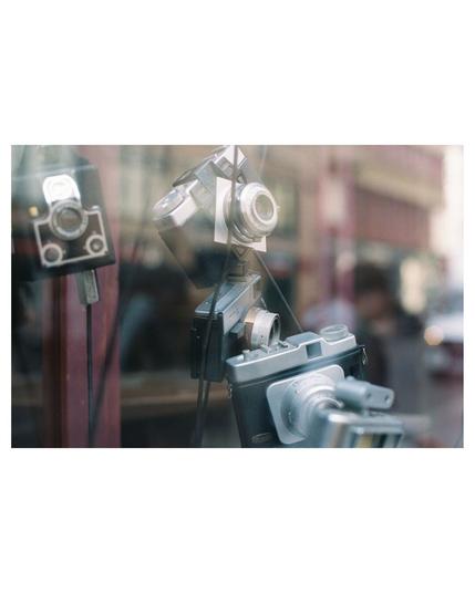vintage cameras hanging in the air of a shop window. the reflections on the window show the blurry contures of city buildings, passing by cars and passerbies