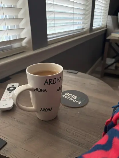 A mug with "AROHA" written on it is on a wooden table beside a remote and coaster, with blinds letting in light from a window.