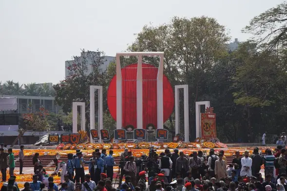 The image depicts a public gathering with a large monument featuring a prominent red circle, which symbolizes the martyrdom of students who fought for the Bengali language. Surrounding the monument are floral arrangements and decorations, typical of a celebration or commemoration. The setting suggests it is likely an event related to language or cultural identity, possibly in Bangladesh or among Bengali communities. There's a large crowd present, indicating a significant communal observance.

Image Credits: Wikimedia / Subarashitours / CC BY-SA 4.0