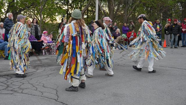 Six morris dancers in rag coats dance a stick dance. An audience looks on from behind.