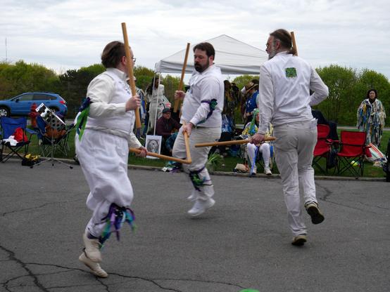 Three morris dancers in white kit dance a stick dance. Other dancers look on from behind.