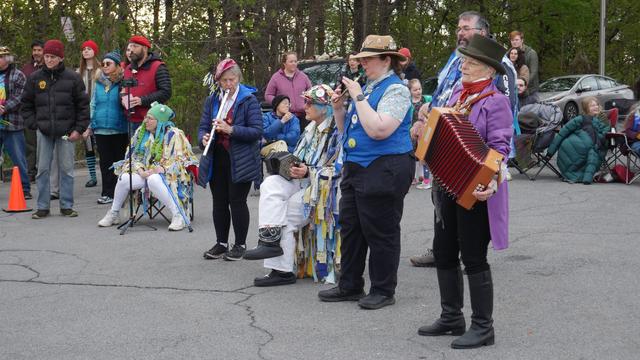 A group of musicians play pennywhistle, concertina, melodeon, and drum. An audience is watching the out of frame dancing.