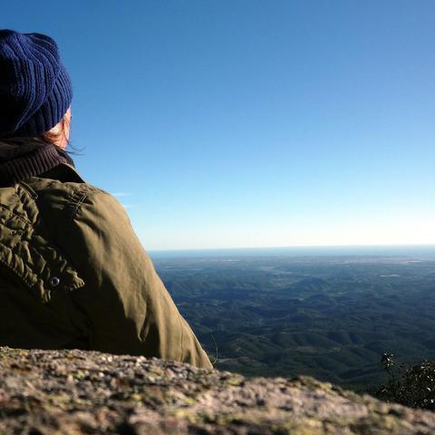 Person mit blauer Mütze und grüner Jacke sitzt auf einem Felsen und blickt auf ein weites, bewaldetes Hügelland der Serra de Monchique in der Algarve, mit dem Atlantik am Horizont unter klarem, blauem Himmel.