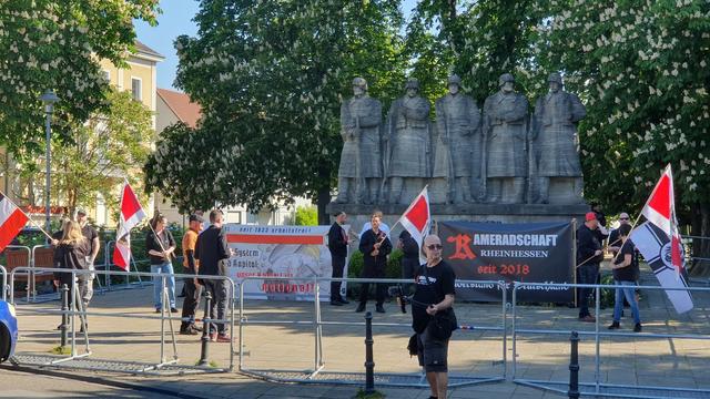 Eine Gruppe Nazis mit Bannern und Fahnen stehen vor dem 118er Kriegerdenkmal in Worms in einem abgegitterten Versammlungsbereich.