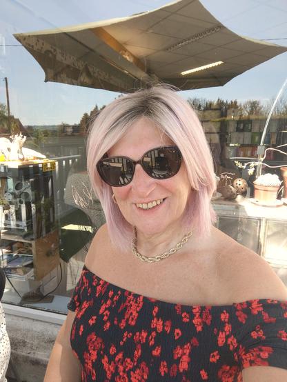 A pink haired woman sitting outside at a cafe, wearing bardot dress in black with small white flowers, a gold chain necklace, gold hoop earrings and sunglasses.
There is a large umbrella and seats reflected in the window behind me.