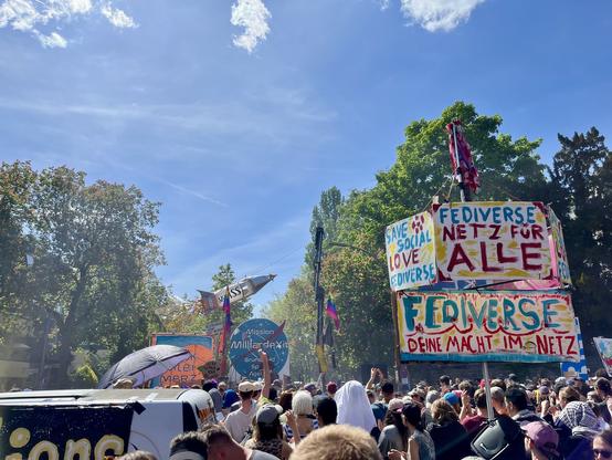 Viele Menschen schicken eine gebastelte Rakete in den Himmel; viele bunter Schilder mit Aufschriften wie „fediverse. Deine Macht im Netz“; blauer Himmel und viele Bäume