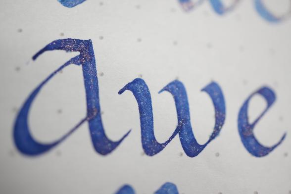 "Awe" in periwinkle ink with gold glitter, written in Carolingian minuscule