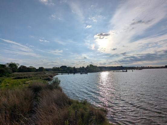 Photo of a calm morning sunrise over Harris Creek sun rising low to the left behind a cloud bedecked sky. Marsh is green with riotous new spring growth. Creek water is rippled and reflects the sky, sun and clouds.