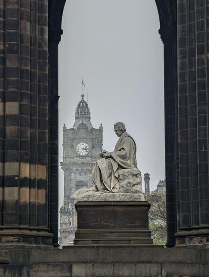 In the foreground, a sculpture of a man seated and facing to the left between two dark stone columns. In the background, a clocktower to the left of the seated figure so it appears he is facing it.