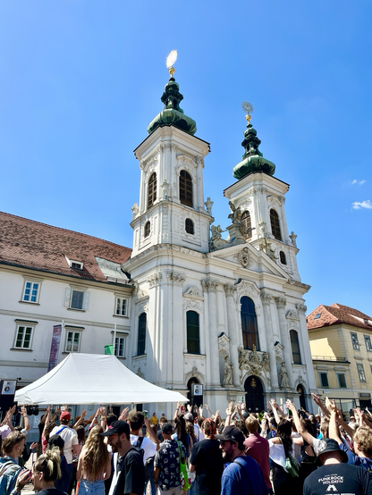 Städtischer Platz mit vielen Personen vor einer Bühne mit Blick auf die Mariahilfer Kirche