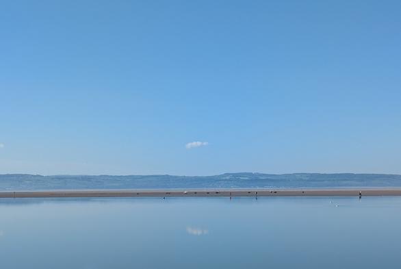 A tranquil seascape features a calm body of water reflecting the clear blue sky and single cloud, with distant hills and tiny figures walking along the lake wall.