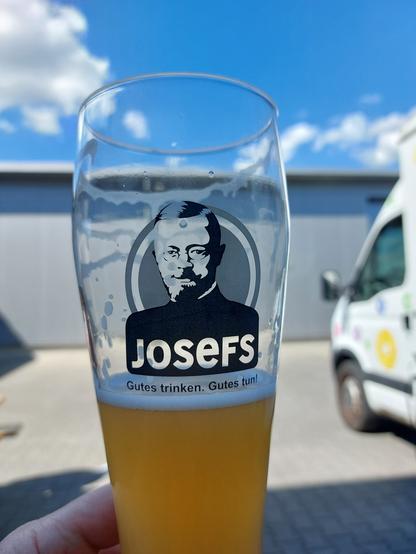 Halb gefülltes Weizenglas mit einem Aufdruck. Logo eines Mannes mit Spitzbart. Beschriftung: Josef- Gutes trinken. Gutes tun.