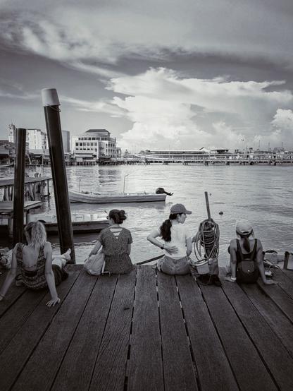 Several tourists sit at the edge of a shady pier resting and enjoying the light breeze, in the distance large cumulonimbus form over the mainland and slowly drift south over the strait towards Penang