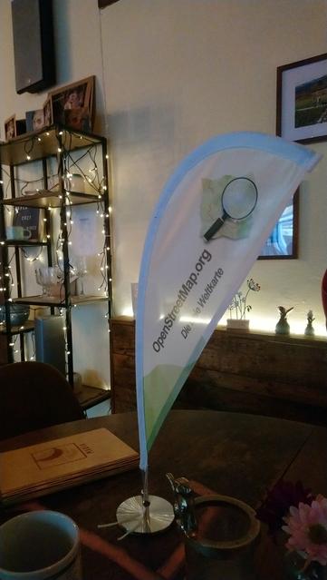 Eine kleine Fahne mit OpenStreetMap-Logo steht auf einem Tisch in einem Café. Es ist abendlichen stimmungsvoll eingerichtet und beleuchtet
