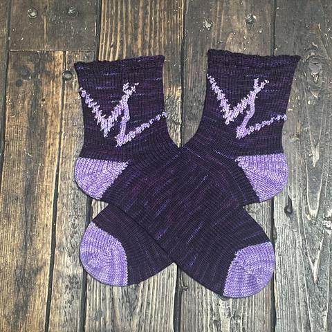 A pair of hand knit socks are laid flat on a wooden surface. They are dark purple with lighter purple toes, heels, and knitted interlocking VL pattern on the ankle. #loomknitting #iwtv #iwtvart