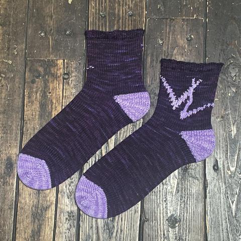 A pair of hand knit socks are laid flat on a wooden surface. They are dark purple with lighter purple toes, heels, and knitted interlocking VL pattern on the ankle. You can see that the VL is only on one side of the ankle, with the other sode plain.